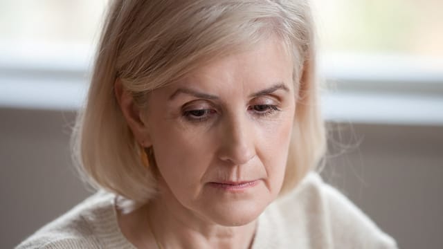 Close-up of a middle-aged woman with light blonde hair and fair skin, looking down with a thoughtful or serious expression, wearing a cream-colored sweater.
