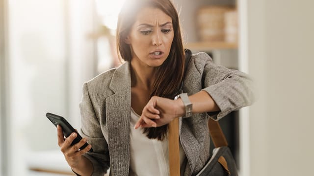 A woman holding a smartphone looks worriedly at her wristwatch, appearing constantly busy or late. Dressed in business attire with a bag over her shoulder, she stands indoors in a well-lit room, reflecting the psychology of self-avoidance.