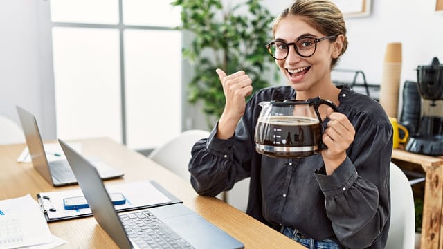 A smiling person wearing glasses sits at a desk with laptops, holding a large coffee pot in one hand and giving a thumbs up with the other. The workspace has plants and coffee supplies in the background.