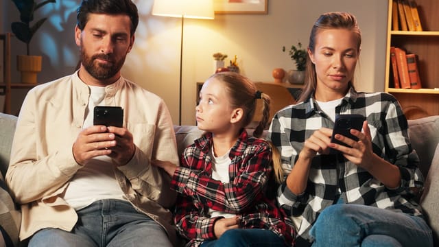 A man and woman sit on a couch using their smartphones, while a young girl between them looks at the man, appearing to seek attention. The setting is a cozy living room.
