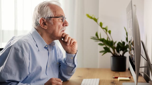 An older boomer dad with gray hair and glasses sits at a desk, looking intently at a computer monitor—perhaps scrolling through Facebook. He rests his chin on his hand, beside a potted plant and keyboard.