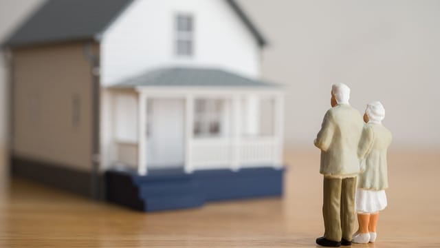 Two small figurines of an elderly couple stand together facing a model house, capturing the deep place attachment and emotions that come with grief over selling a home. The softly blurred house in the background underscores their poignant farewell.