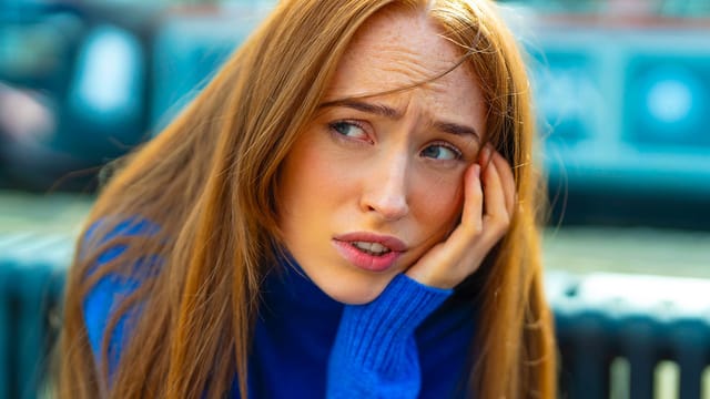 A woman with long red hair wearing a blue sweater looks worried, resting her face on her hand. The background is blurred, capturing the quiet weight eldest daughters often feel as they navigate complex family roles.
