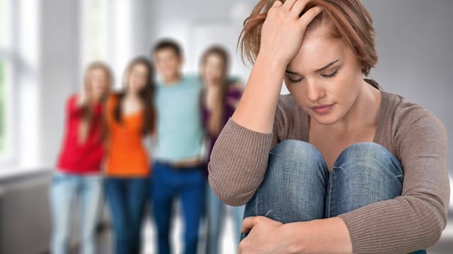 A young woman with rare traits sits alone, looking upset with her head resting on her hand, while a group of four people stands together in the blurred background.