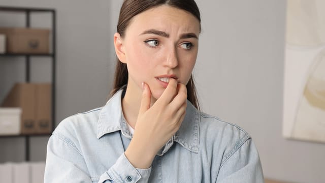 A young woman in a light denim shirt looks to the side with a worried expression, biting her fingernail and furrowing her brow—perhaps weighed down by shrinking happiness after another comparison with others. Shelves of boxes and books blur in the background.