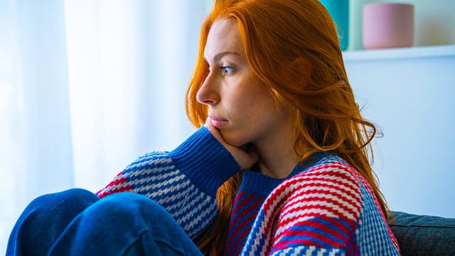 A woman with long red hair wearing a colorful striped sweater sits indoors, resting her chin on her hand and gazing thoughtfully to the side, capturing a quiet moment of solitude as natural light streams through the window in the background.
