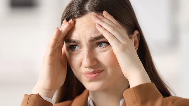 A woman with long brown hair holds her head with both hands and appears stressed or worried, with a furrowed brow and a concerned expression, capturing the overwhelming pressures that often come with adult life.
