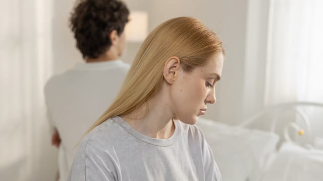 A woman with blonde hair looks down sadly while a man with dark curly hair sits behind her, facing away. Both appear upset in a bright bedroom, highlighting relationship challenges and moments of marriage loneliness.