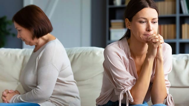 Two women sit on a couch facing away from each other, both looking upset and thoughtful, suggesting family conflict or tension in their parent relationships. One woman rests her chin on her hands, while the other looks down, capturing the complexities often present between adult children and their parents.