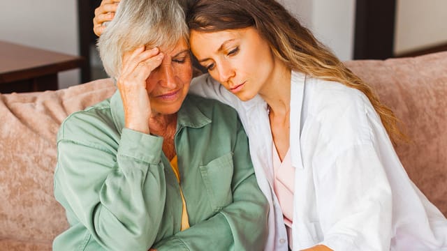 An older woman looks distressed while a younger woman sits beside her, offering comfort with an arm around her shoulders. They sit close together on a couch, capturing a heartfelt moment of caregiving and support as they navigate the challenges of parent aging together.