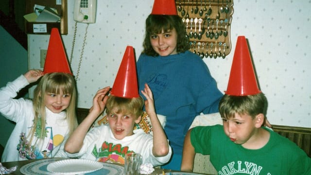 Four children, wearing red party hats and smiles, gather around a table set for celebration—an example of the ordinary luxuries cherished by younger generations. Behind them, a wall with keys adds a quirky touch reminiscent of the 80s and 90s.