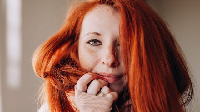 A person with long, vibrant red hair and freckles gazes softly at the camera, holding a strand of hair and wearing a light-colored sweater. The softly blurred background hints at their journey of personal growth.