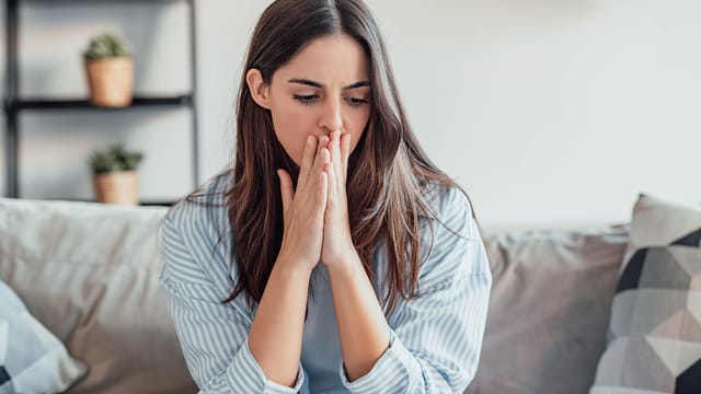A young woman sits on a couch indoors, pressing her hands to her mouth and looking down with a worried or thoughtful expression, perhaps contemplating the emotional risk that often comes with maintaining high standards in relationships. Potted plants and shelves are visible in the background.