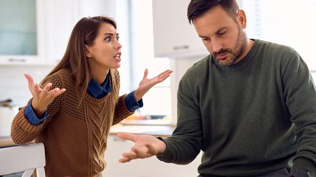 A woman appears upset, raising her hands and speaking to a man who sits with his head down, looking frustrated. They are indoors, possibly in a kitchen, both wearing sweaters—clear signs in relationships of reaching breaking point.