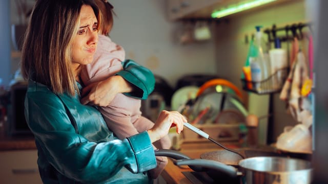 A tired woman in pajamas holds a child on her hip while stirring food in a cluttered kitchen, embodying the psychological effects of motherhood identity as she looks stressed and overwhelmed.