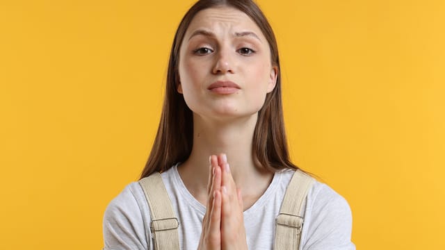 A young woman with long brown hair, wearing a light shirt, stands against a yellow background, holding her hands together in a pleading gesture—capturing the psychology of over-apologizing with her hopeful expression.