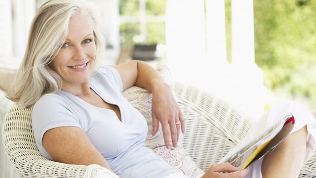 A woman with gray hair sits on a white wicker chair outdoors, smiling and holding a magazine. Radiating happiness in her 60s and 70s, she looks relaxed in her light blue top, surrounded by greenery and sunlight.