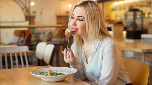 A woman with long blonde hair, wearing a white blouse, sits at a wooden table in a restaurant, enjoying a salad. The softly lit, cozy background sets the scene as she unwinds and practices healthy coping mechanisms during her meal.