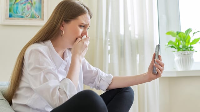 A woman with long hair sits indoors, holding a smartphone and looking at the screen with a worried expression, covering her mouth with her hand. Light streams through sheer curtains behind her, hinting at a human tragedy unfolding through psychology news.