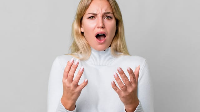 A woman with long blonde hair wearing a white sweater looks surprised or upset, possibly experiencing decision fatigue, with her mouth open and hands raised in front of her against a plain light background.