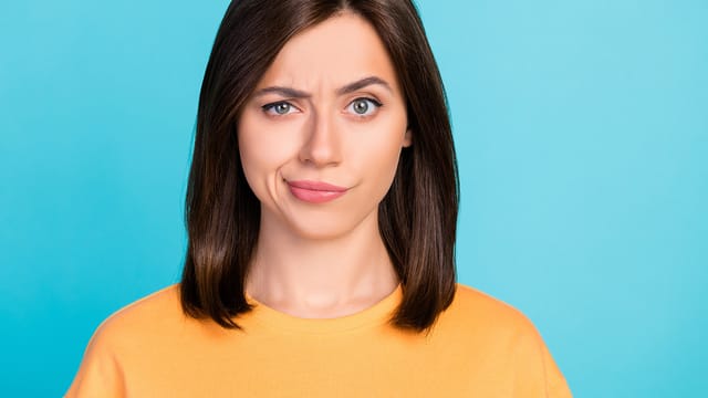 A woman with straight brown hair in a yellow shirt stands against a blue background, raising one eyebrow and smirking, as if questioning manipulation or displaying a strong sense of self through her skeptical expression.