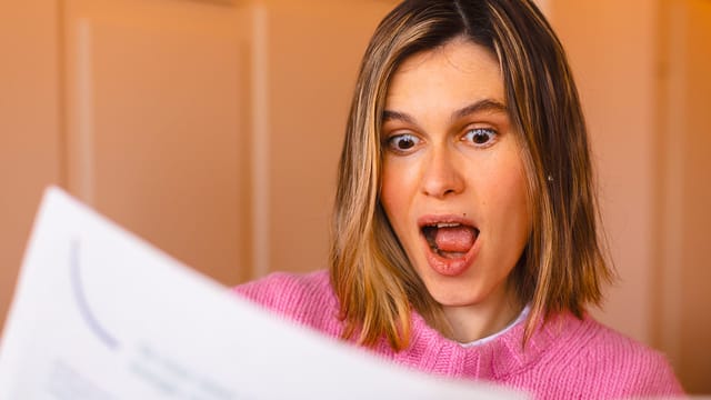 A woman with shoulder-length blonde hair, wearing a pink sweater, looks surprised and excited while reading a document about psychology and horoscopes held in front of her.