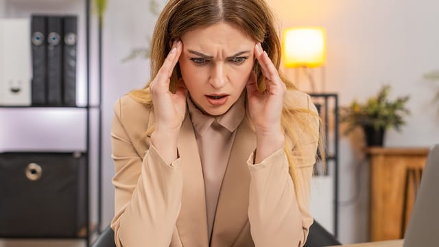 A woman sits at a desk, holding her temples in frustration—a clear sign of millennial exhaustion or burnout—as she stares at her laptop. Office shelves and a lit lamp are visible in the background.