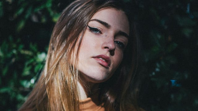 A young woman with long, light brown hair and blue eyes looks slightly upward, standing against a background of dark green foliage. She wears a neutral expression and a light brown top, as if lost in thought about beliefs and actions. Sunlight highlights her face.
