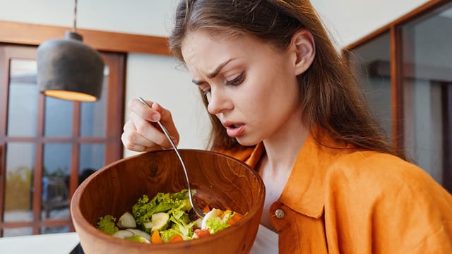 A young woman with long brown hair in an orange shirt looks unhappily at a bowl of salad, holding a fork and appearing displeased or skeptical about sticking to her diet.