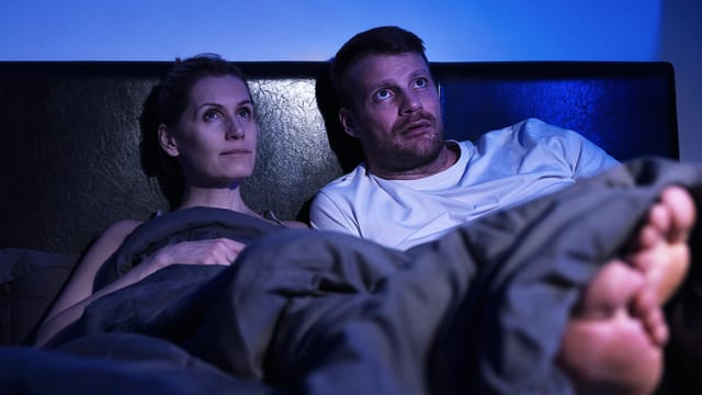 A couple sits up in bed under gray blankets, watching with focused expressions. The dimly lit room glows blue as they follow subtitles intently, lost in the story unfolding on their screen.