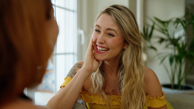 A smiling woman with long blonde hair and a yellow off-shoulder top rests her hand on her face while talking to another person indoors, showing signs of confidence as she appears happy, engaged, and secure in a room filled with sunlight.