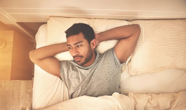 Above view of depressed man, lying in bed and staring. Sad tired male waking up late in morning before starting the day early. Stressed exhausted young guy thinking about problems and difficulties