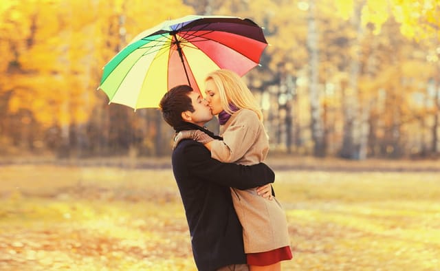 Happy loving young couple kissing and hugging under colorful umbrella in autumn park on warm sunny day