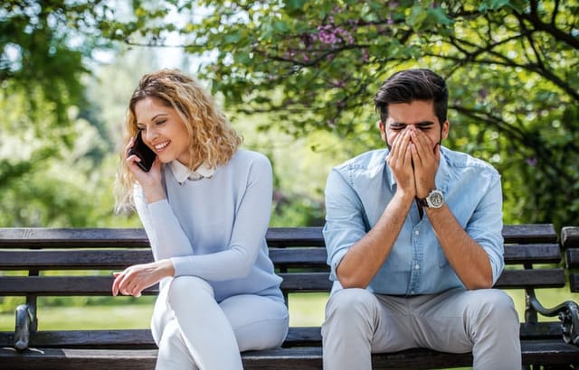 Cheating, jealous, obsession, possession. Young woman talking on smartphone while angry boyfriend sitting next to her on the park bench. Bad love relationships