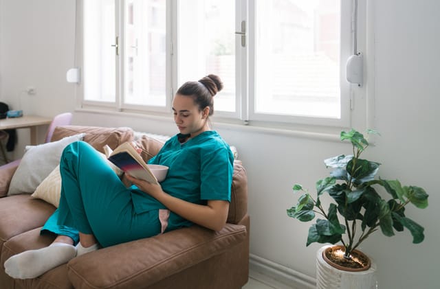 Photo of a female healthcare professional still in her medical scrubs, eating a meal and reading a book in her living room as a way of relaxation after a job in a hospital