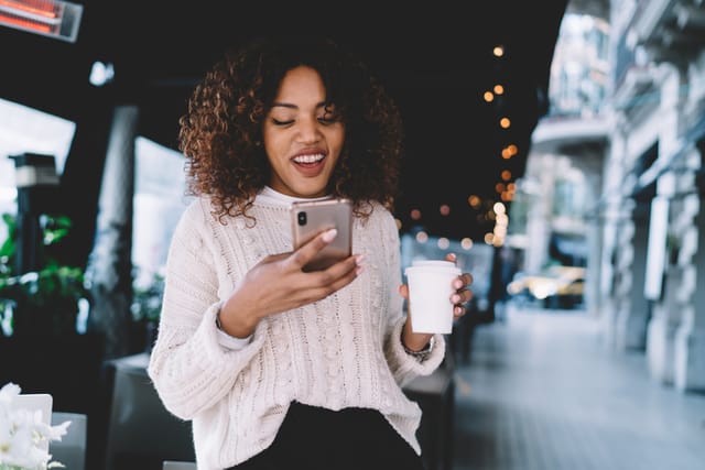smiling black woman texting with coffee outdoors