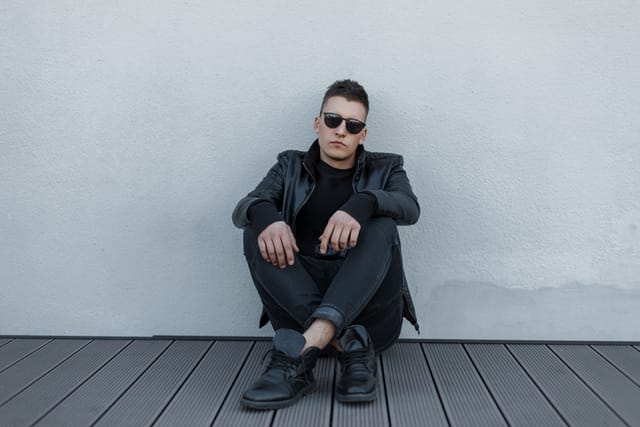 Attractive young hipster man in black sunglasses, black leather jacket, stylish sweatshirt and trendy jeans sits on a wooden floor near a gray wall. Handsome guy