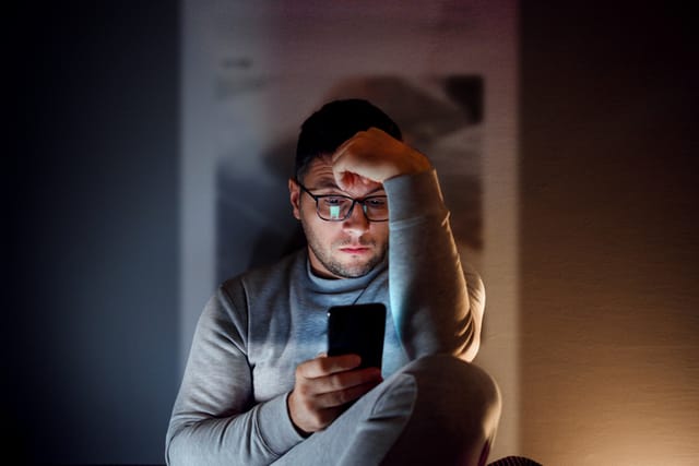 A 35-year-old guy is sitting in the dark room and using his smartphone.