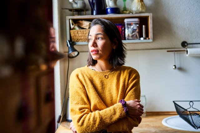 Young woman in the kitchen looking away. Thoughtful woman with her arms crossed looking outside the window.