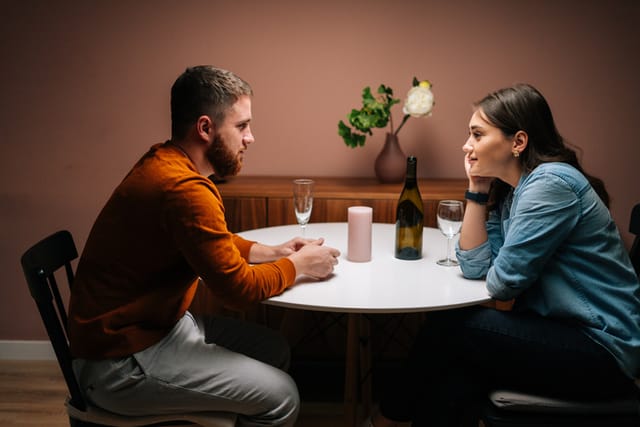 Side view of happy young couple enjoying talking and sitting together at dinner table in cozy dark living room.