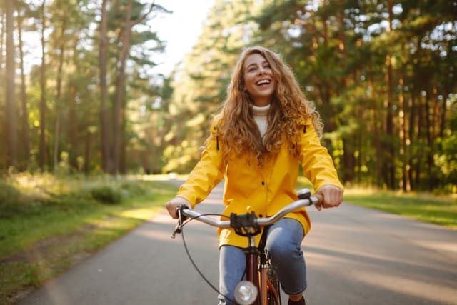 A smiling female tourist in a yellow coat enjoys the weather in the autumn park while riding a bicycle. Autumn fashion. Concept of relaxation, nature.