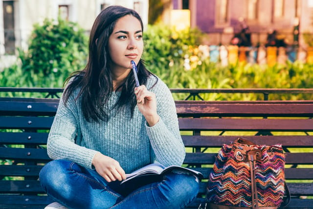 Pensive woman thinking and sitting on bench outdoors. Beautiful young lady making notes and sitting with her legs crossed, city view in background. Urban lifestyle and education concept. Front view.