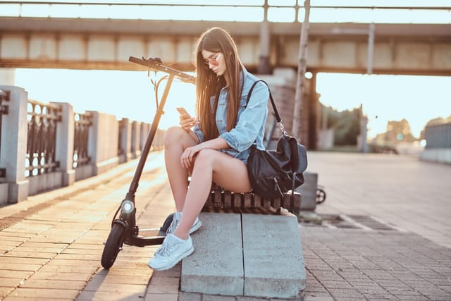 Young attractive woman is chatting by mobile phone while sitting on the bench with her scooter.