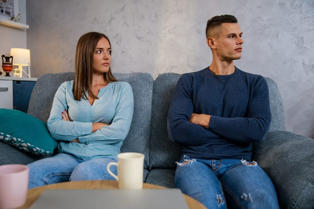 Front view of angry man and woman sitting on the sofa, with arms crossed, after they had a fight due to relationship difficulties. Young woman is looking at her boyfriend, who is looking away.