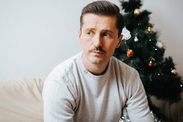 Portrait of upset, sad man sitting on sofa near decorated Christmas tree 