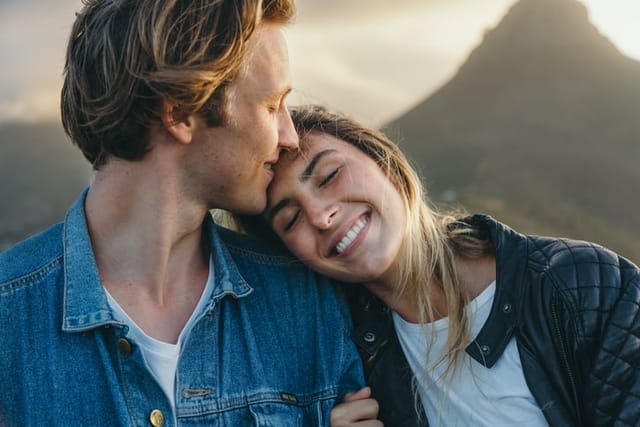 Close-up of smiling beautiful young girlfriend with eyes closed resting head on boyfriend' shoulder against mountain at sunset