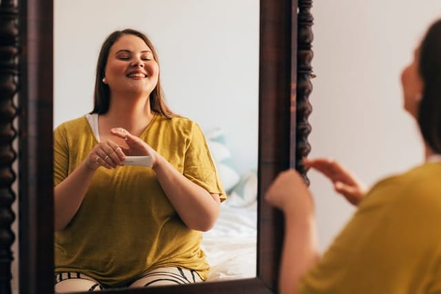 Beautiful Caucasian plus size woman in her bedroom, looking at herself in a big mirror, applying her morning cosmetics.