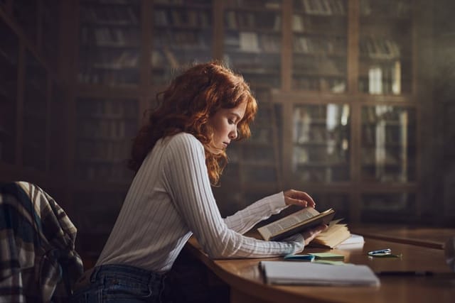 Redhead female student reading a book in library.