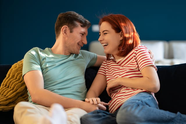 Overjoyed woman sincerely laughing with smiling boyfriend man holding hands, sit on couch at home