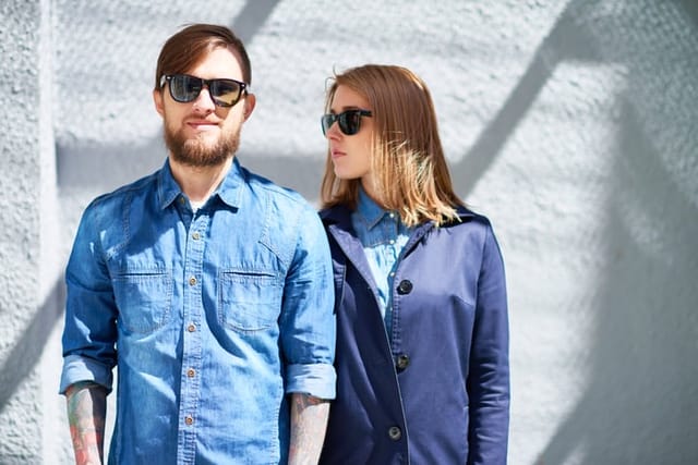 Bearded man in sunglasses looking at camera, young woman in coat standing behind him and looking away
